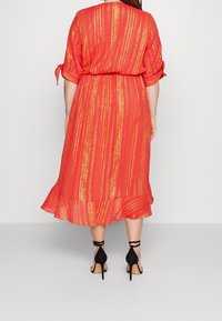 Rotes Kleid mit goldenen Streifen, fließendem Stoff, tailliertem Bund und Rüsche am Saum. Getragen mit schwarzen High-Heels.