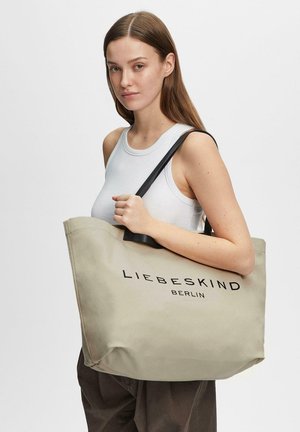 Junge Frau trägt ein weißes Tanktop und hält eine große beige Canvas-Tragetasche mit schwarzem "Liebeskind Berlin"-Logo auf dem Träger und dem Taschenkörper.