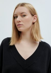 Schwarzer gestrickter Pullover mit tiefem V-Ausschnitt, geripptem Kragen und strukturiertem Stoff. Das Modell hat schulterlanges, glattes, blondes Haar und ein neutrales Make-up.