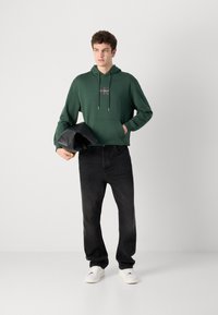 Felpa verde con tasca frontale e logo, abbinata a jeans scuri e sneaker bianchi. La felpa presenta un cordino e una texture morbida.