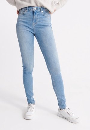Person iført lyseblå skinny jeans, hvide sneakers og en cremefarvet sweater, stående med hænderne i lommerne på en hvid baggrund.