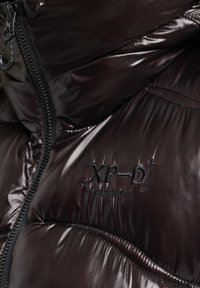 Brązowa kurtka puchowa o błyszczącej, pikowanej fakturze. Posiada wysoki kołnierz oraz czarne haftowane logo "XP-D Superdry" na klatce piersiowej. Zamek błyskawiczny.