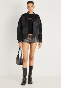 Νεαρή γυναίκα που φοράει μαύρο μπουφάν τύπου bomber, μαύρο crop top, μεταλλικές σορτς, καφέ σκούρες μπότες μέχρι το γόνατο και κρατάει μια μαύρη τσάντα.