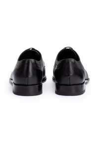 Een paar zwarte leren nette schoenen, gezien van achteren op een witte achtergrond, met de nadruk op de lage hakken en het gladde oppervlak.