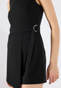 Robe noire sans manches avec une large ceinture et un détail de boucle circulaire en argent. Tissu lisse, coupe ajustée et fente latérale pour un look moderne.
