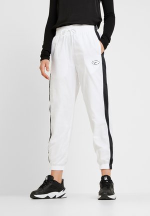 Osoba ubrana w białe spodnie dresowe Nike z czarnymi paskami po bokach i czarne buty sportowe Nike, stojąca na jednolitym jasnym tle.