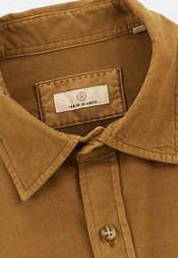 Chemise en velours côtelé marron avec un col pointu, dotée d'une étiquette portant la mention "SERGE BLANCO" et d'un détail de bouton bien en vedette.