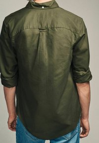 Camicia verde oliva con maniche arrotolate, colletto e dettaglio con piega sul retro, realizzata in tessuto liscio; abbinata a jeans blu.