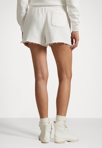 Weiße Baumwollshorts mit fransigem Saum und Gesäßtasche, kombiniert mit eng anliegenden weißen Stiefeletten und gerippten Socken. Einfaches und lässiges Design.