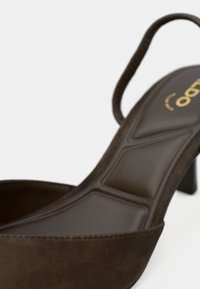 Chaussure à talon haut en daim marron avec une semelle intérieure rembourrée et une fine bride à la cheville sur un fond uni.