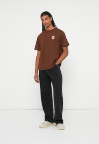 Brun bomuld t-shirt med et lille grafik på venstre bryst, parret med sorte jeans og off-white sneakers på en neutral baggrund.