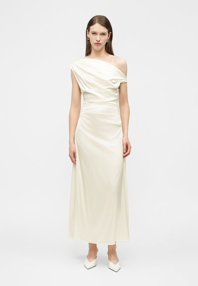 Γυναίκα που φοράει ένα μακρύ, λευκό σατέν φόρεμα με έναν ώμο, με ντραπαρισμένο κορσάζ και λευκά μυτερά τακούνια, στέκεται μπροστά σε απλό φόντο.