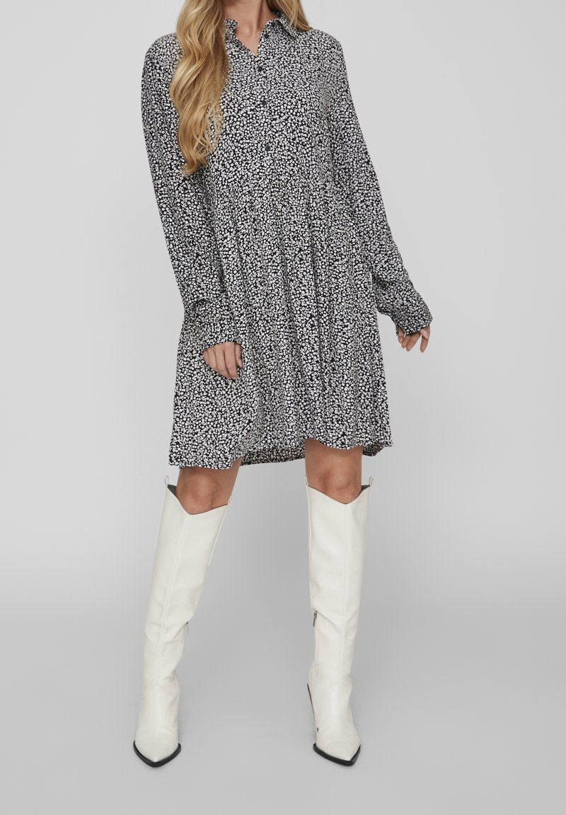 Vestito in stampa floreale bianco e nero con colletto, maniche lunghe e silhouette svasata, abbinato a stivali bianchi al ginocchio.