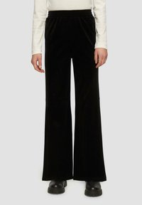 Persoon in zwarte wijde broek, zwarte chunky schoenen en een wit langemouwtop, staande tegen een effen grijze achtergrond.