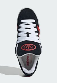 Sneaker in suede nero con lacci bianchi, un accento rosso e un logo sulla linguetta. L'interno è foderato con materiale tessile bianco.