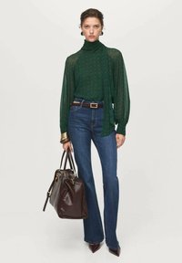 Blusa verde con estampado y cuello alto, combinada con unos jeans acampanados azules y un bolso de cuero marrón. Pulsera dorada como acento en la muñeca.