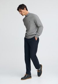 Pull gris à col montant, pantalon foncé et chaussures noires avec lacets marron. Le pull comporte un petit emblème sur le côté gauche de la poitrine.