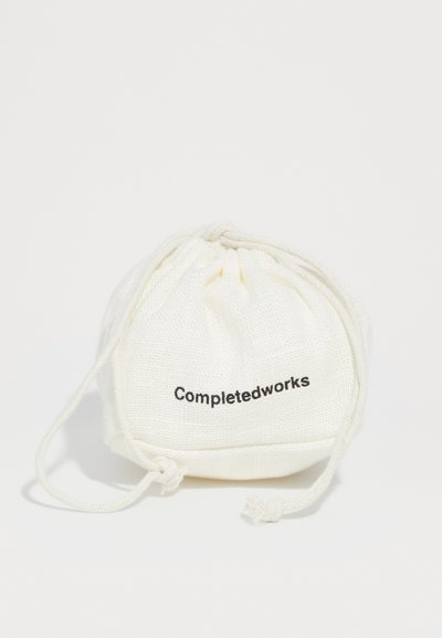 Kreemi tekstuuriga off-white kanga tõmbluku kott, millel on sõlmega nöörid ja mille esiküljel on musta värviga trükitud "Completedworks".