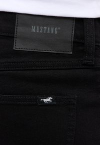 Schwarze Denim-Jeans mit einem schwarzen Lederetikett, das "MUSTANG" zeigt, und einem kleinen weißen Pferdelogo auf der vorderen Tasche.