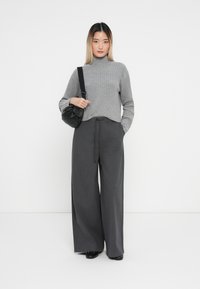 Grauer gerippter Rollkragenpullover kombiniert mit hoch taillierten, weit geschnittenen, anthrazitfarbenen Hosen. Eine schwarze Umhängetasche setzt Akzente. Sanfte Textur durchgehend.