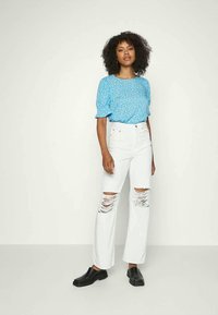 Mulher com cabelo encaracolado vestindo uma blusa floral azul, jeans brancos rasgados e sapatos pretos de slip-on, encostada a um fundo liso.