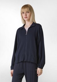 Giacca zip-up navy in morbido tessuto con colletto rialzato, polsini elasticizzati e vestibilità comoda, abbinata a pantaloni scuri coordinati.