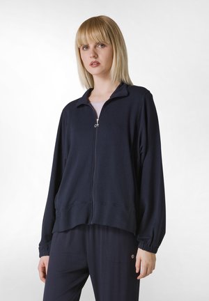Giacca zip-up navy in morbido tessuto con colletto rialzato, polsini elasticizzati e vestibilità comoda, abbinata a pantaloni scuri coordinati.