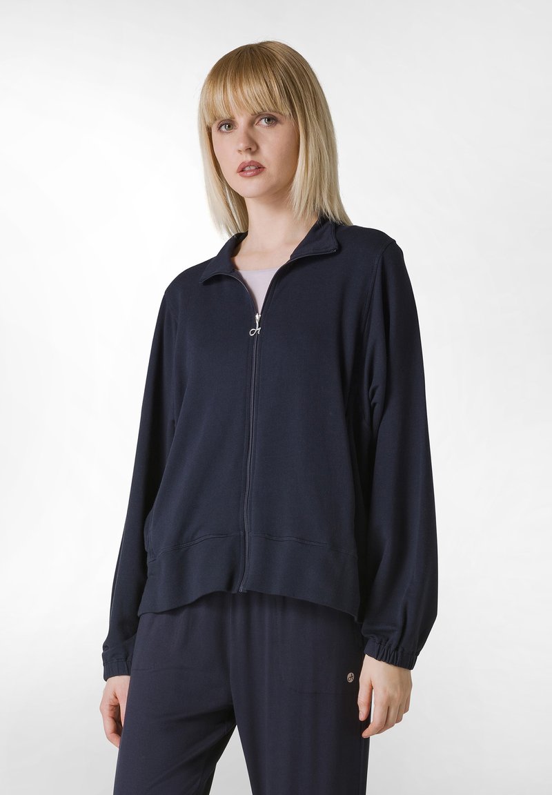 Giacca zip-up navy in morbido tessuto con colletto rialzato, polsini elasticizzati e vestibilità comoda, abbinata a pantaloni scuri coordinati.