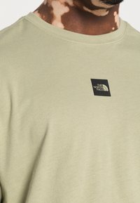 Ljusgrön bomulls-T-shirt med rund halsringning och en svart fyrkantig logo på bröstet. Mjuk textur med slät yta.