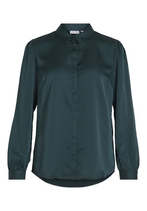 Femme portant un chemisier en satin vert foncé, un pantalon noir à jambes larges et des bottines noires, debout les mains dans les poches.