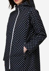 Musta sateenjakku, jossa on huppu, vetoketjullinen etuosa ja avoimet taskut. Ominaisuuksina valkoiset pilkut ja teksturoitu kangas.