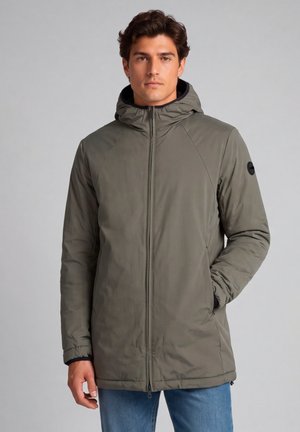 Uomo che indossa una giacca invernale verde con cappuccio, chiusa con la cerniera, con una mano in tasca e jeans blu su uno sfondo grigio uniforme.