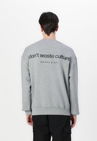 Sudadera gris con mangas largas, puños y dobladillo acanalados. Presenta un gran texto negro en la parte posterior: "no desperdicies cultura MAISON D'ART."