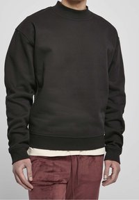Sudadera negra con cuello alto, puños y dobladillo acanalados. Tela suave y texturizada, con un corte holgado. Llevada sobre una camiseta de color claro.