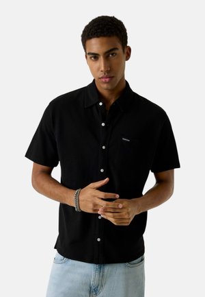 Giovane uomo con indosso una camicia nera a maniche corte con bottoni e jeans azzurri chiari, che posa con le mani giunte davanti a uno sfondo bianco.