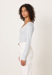 Svijetlosivi top dugih rukava s širokim V-izrezom, uparen s visokim strukom bijelih traperica i suptilnim detaljima šavova. Ton kože vidljiv na ruci.