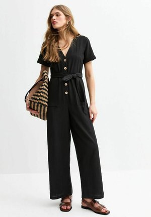 Black jumpsuit with short sleeves, deep V-neck, wooden buttons, tied waist, side pockets, paired with a striped bag and brown sandals.