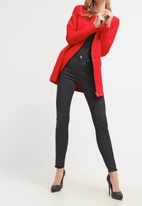 Roter Blazer mit langem Schnitt, Knopfdetails und schwarzen Skinny-Jeans. Das Model trägt schwarze High Heels. Glatte Texturen und leuchtende Farben.
