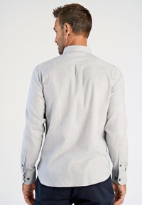 Uomo in piedi, di spalle, con una camicia grigia chiaro a maniche lunghe e pantaloni scuri, su uno sfondo neutro.