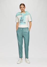 Eine tealfarbene Kunstlederhose mit elastischem Bund, kombiniert mit einem weißen T-Shirt mit Grafik in Blau und einem azurblauen Strickpullover, der drapiert ist.