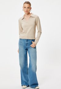 Beżowa bluzka z długim rękawem w kształcie V z kołnierzykiem, połączona z jasnoniebieskimi spodniami typu wide-leg. Gładka tekstura, minimalistyczny design i swobodny wygląd.