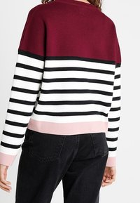 Sweter dziany z bordowym pasem u góry, czarno-białym paskowanym korpusem i różowym obszyciem. Posiada długie rękawy i żeberkowaną fakturę.