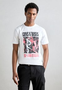 Balta medvilninė marškinėlių su drąsiu grafiniu spaudiniu, kuriame yra juodų ir raudonų elementų, įskaitant tekstą "GUNS N' ROSES" ir "USE YOUR ILLUSION."