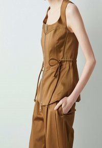 Top marrone con zip frontale, corsetto aderente, vita arricciata e dettaglio a fascia regolabile. Texture liscia, design senza maniche e leggero lucido.