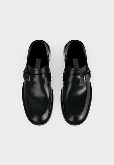 Ζεύγος μαύρα δερμάτινα loafers με πλευρικά αγκράφες και στρογγυλεμένες μύτες σε απλό φωτεινό φόντο.