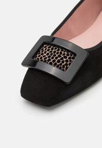 Chaussure plate en daim noir à bout carré, ornée d'une grande boucle noire brillante sur un détail en tissu imprimé léopard. Doublure intérieure rose pâle visible.