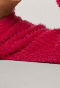 Plyšový, jasně růžový svetr s texturovaným pleteným vzorem, volného střihu a širokými rukávy. Pozadí je jednoduché a neutrální.