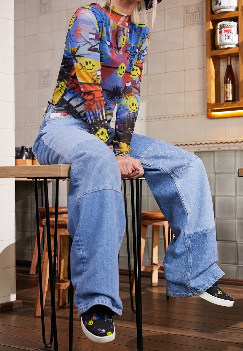 Colorful long-sleeve top featuring smiley face prints and abstract designs paired with wide-leg, light blue denim jeans and patterned shoes.