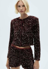 Giacca di paillettes bordeaux con maniche lunghe, scollo tondo e chiusura con bottone, abbinata a pantaloni corti di paillettes coordinati. Texture liscia con dettagli lucidi.