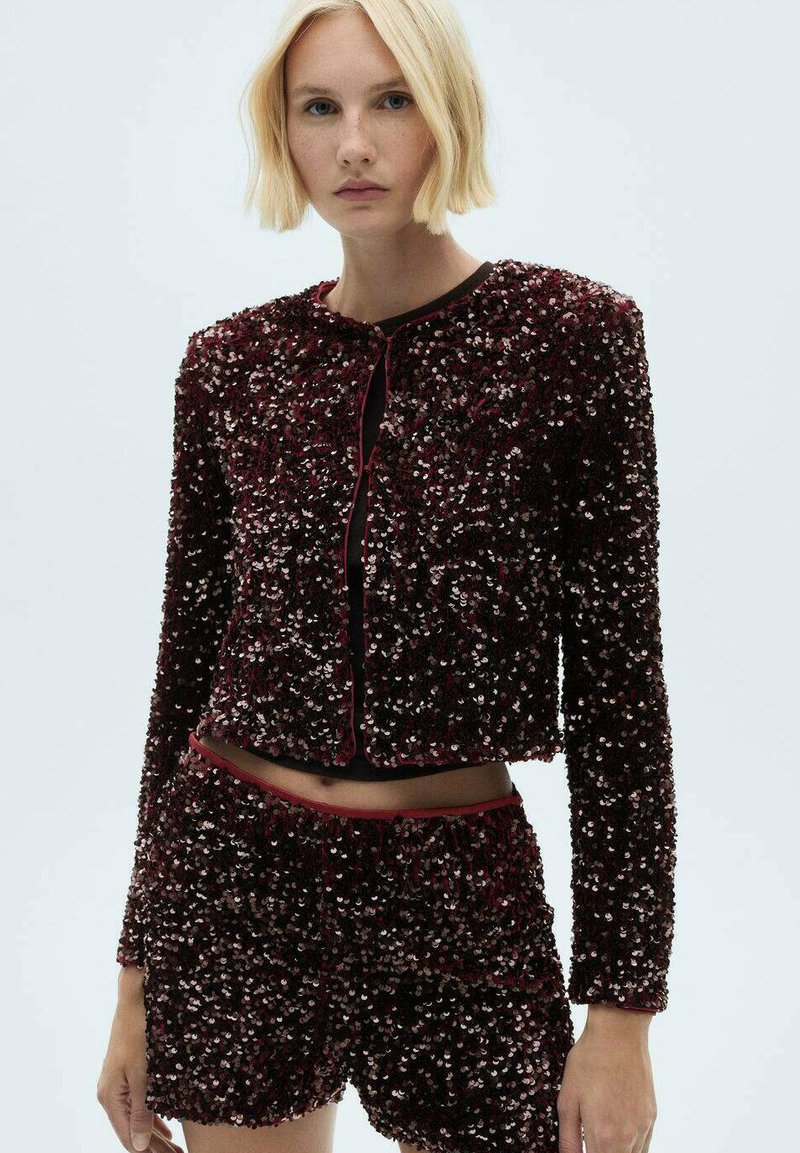 Giacca di paillettes bordeaux con maniche lunghe, scollo tondo e chiusura con bottone, abbinata a pantaloni corti di paillettes coordinati. Texture liscia con dettagli lucidi.